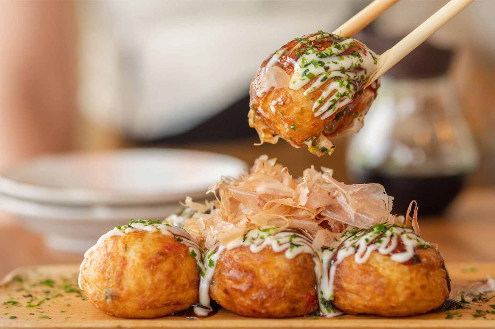 Chopsticks lifting a hot Takoyaki octopus ball from a serving board topped with bonito flakes, dried seaweed, and savory sauce.