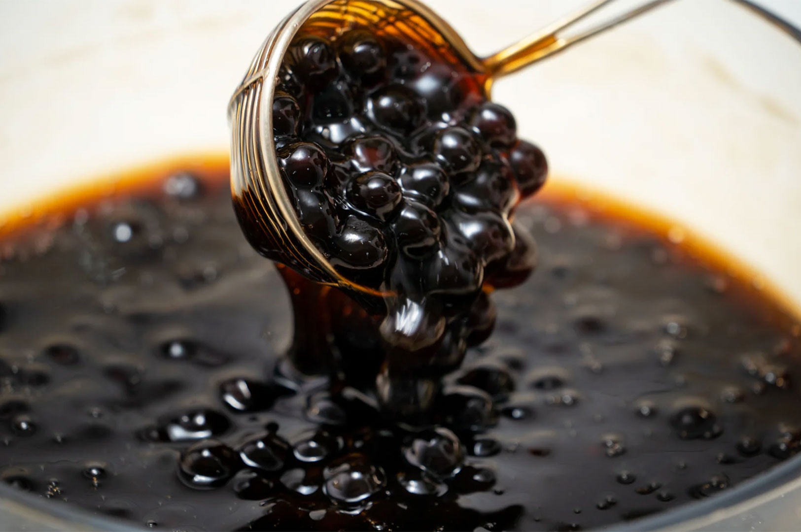 A wire strainer lifting glistening, cooked black tapioca pearls out of a thick, dark brown sugar syrup.