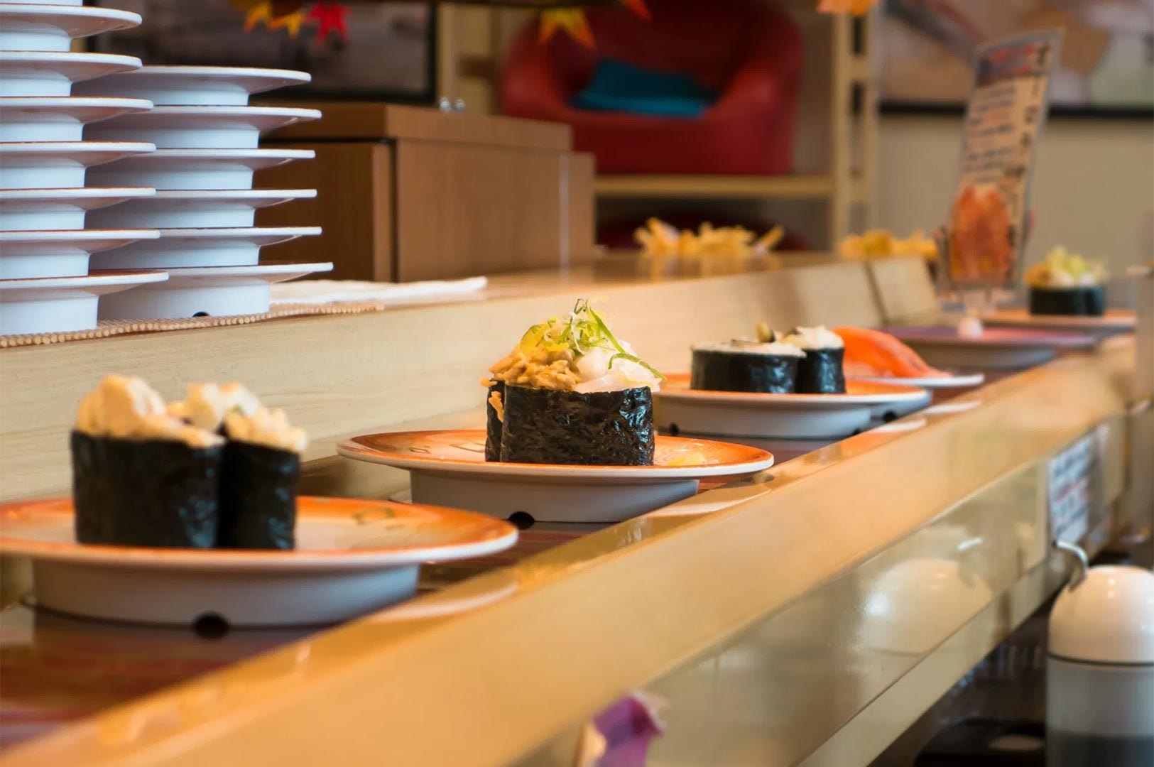 Plates of sushi, including gunkan maki, moving along a conveyor belt at a Kaitenzushi restaurant with stacked plates in the background.