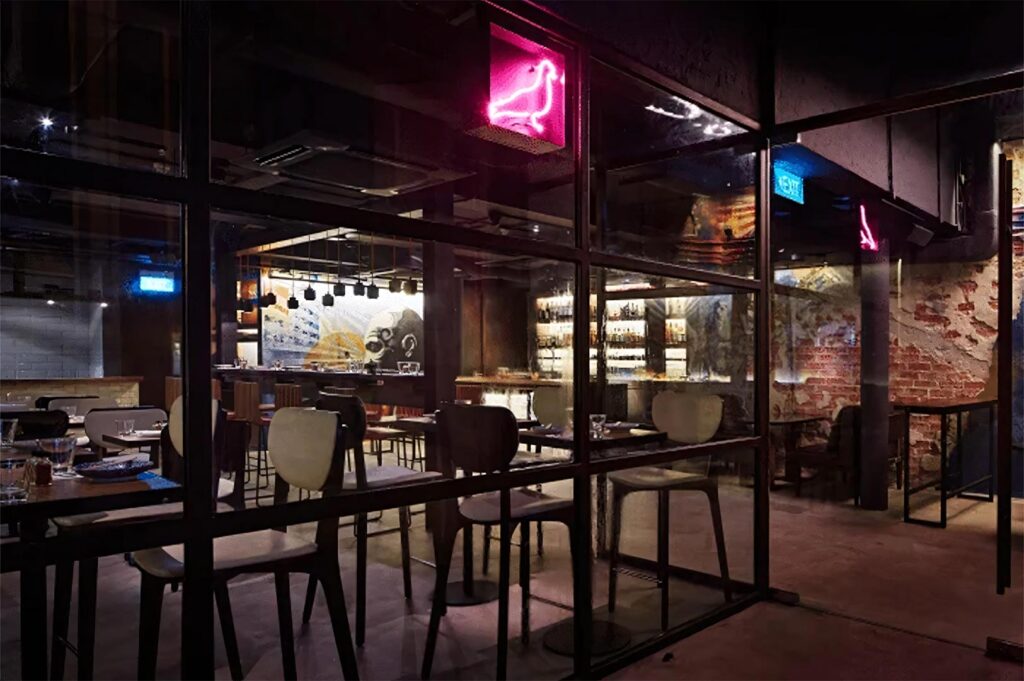 A dark, modern Japanese restaurant interior featuring pink neon pigeon signs, industrial glass partitions, and a well-lit bar in the background.