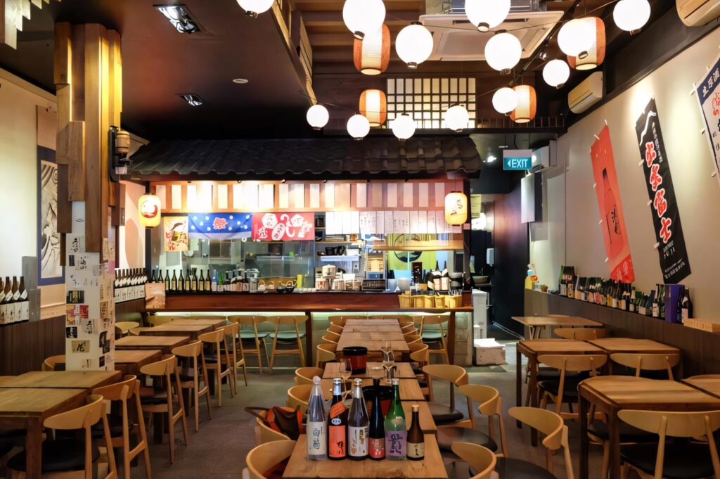 A vibrant Japanese izakaya dining area with wooden tables, paper lantern lighting, and a traditional shingled roof over the kitchen counter.