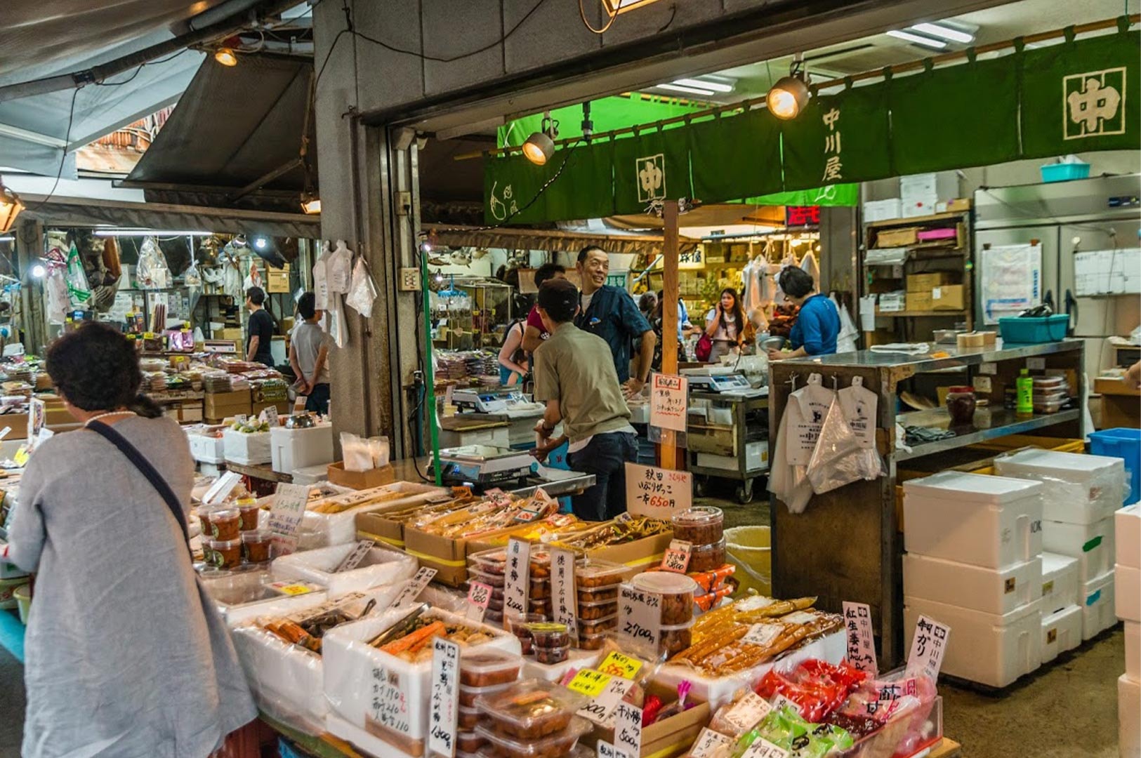 A bustling traditional Japanese market scene featuring vendors selling pickles and dried goods from wooden stalls with green fabric noren curtains.