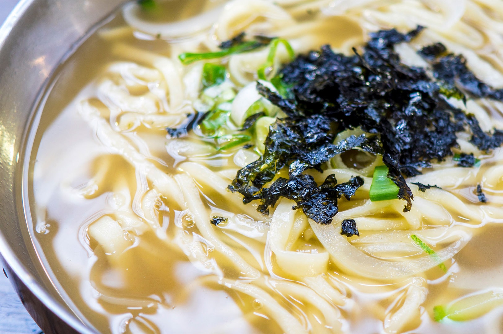 A stainless steel bowl of Kalguksu, Korean knife-cut noodles in a clear broth, topped with shredded seaweed (gim) and chopped green onions.