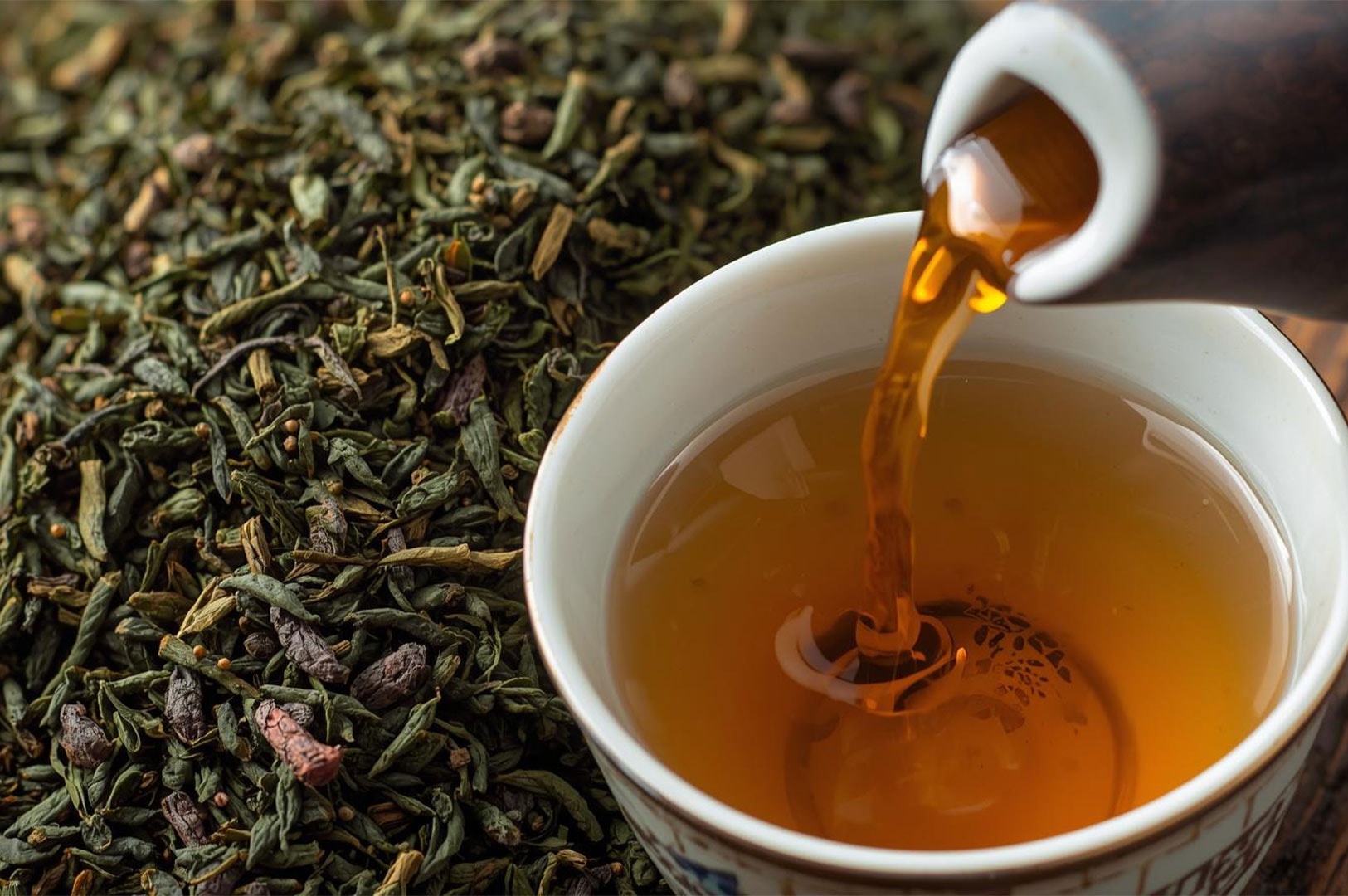 A close-up view of amber-colored tea being poured into a white cup, set against a textured background of loose dried tea leaves and twigs.