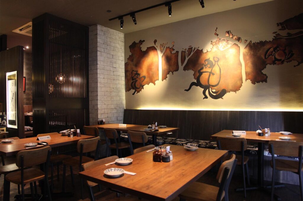 Warmly lit restaurant interior featuring wooden dining tables, dark wood partitions, and a large golden wall mural depicting traditional Japanese folklore.
