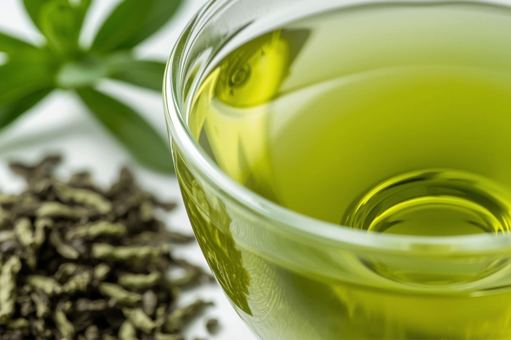 A clear glass cup containing freshly brewed yellow-green tea, sitting next to a pile of dried loose-leaf green tea leaves.