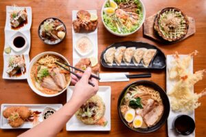 Flat lay of a grand Japanese feast featuring bowls of ramen, crispy tempura shrimp, gyoza dumplings, fried rice, and sushi, with a hand using chopsticks.