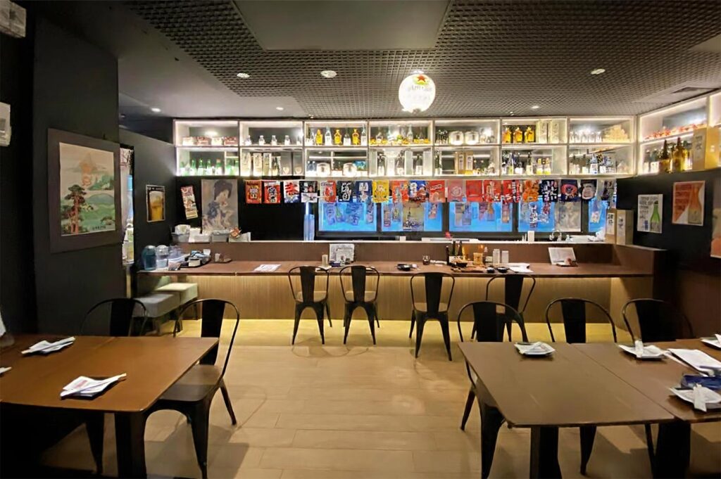A clean, minimalist Japanese bar counter with shelving displaying sake bottles, colorful tapestries, and black industrial-style seating.