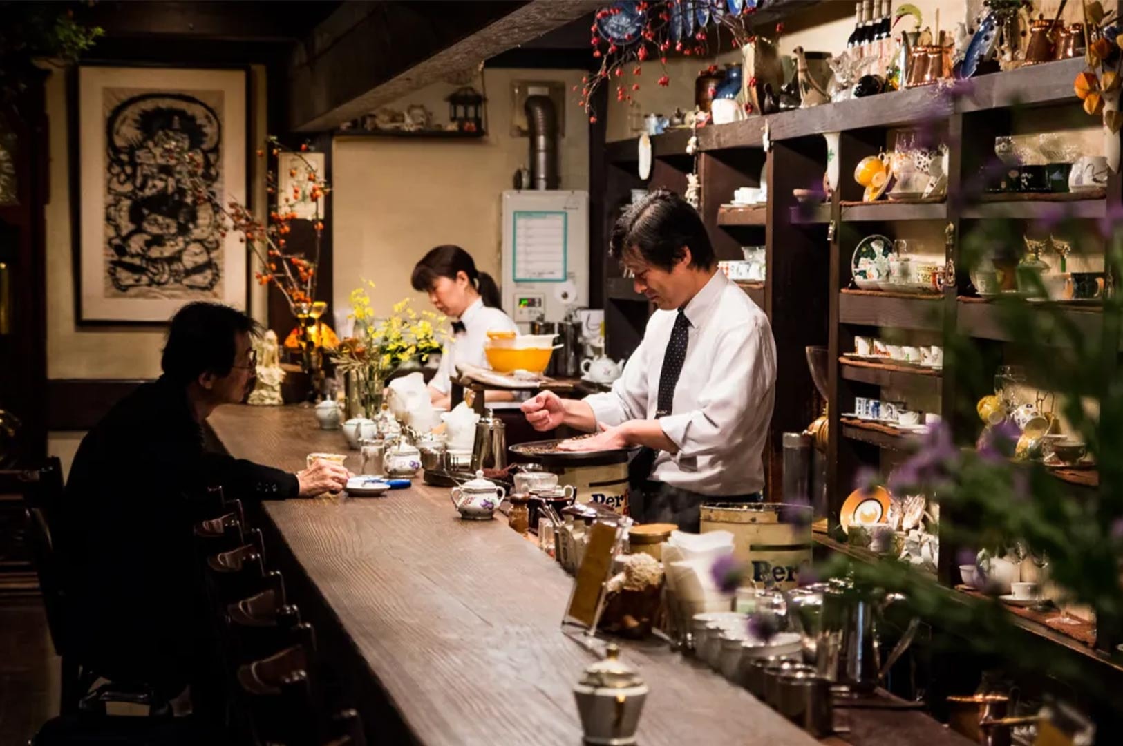 The interior of a retro Japanese Kissaten coffee shop featuring a barista working behind a long wooden counter and a customer seated in a cozy, cluttered atmosphere.