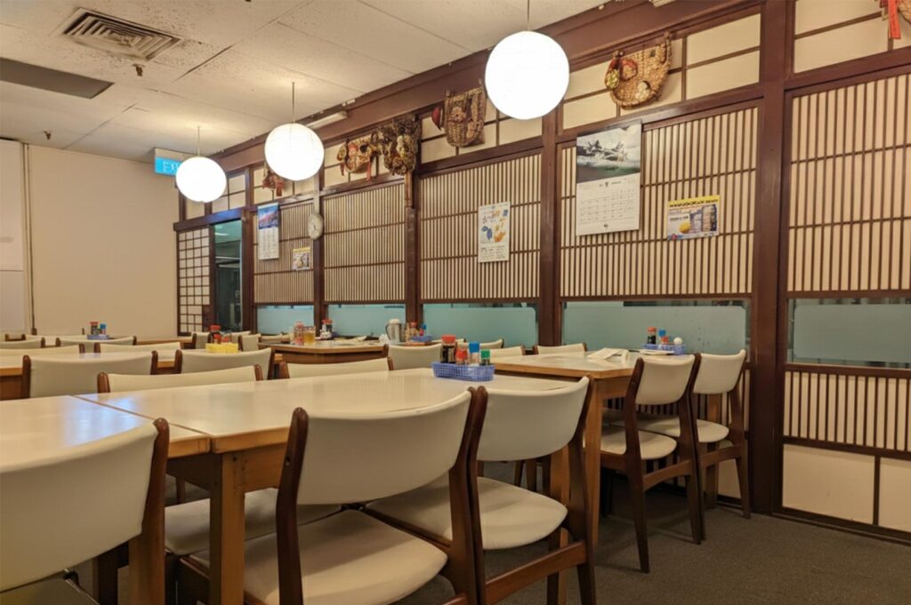 A bright, traditional Japanese restaurant interior with white chairs, wooden tables, round paper lanterns, and classic shoji-style wall panels.