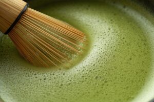 A close-up view of a traditional bamboo whisk (chasen) creating a rich foam while stirring vibrant green matcha tea in a bowl.