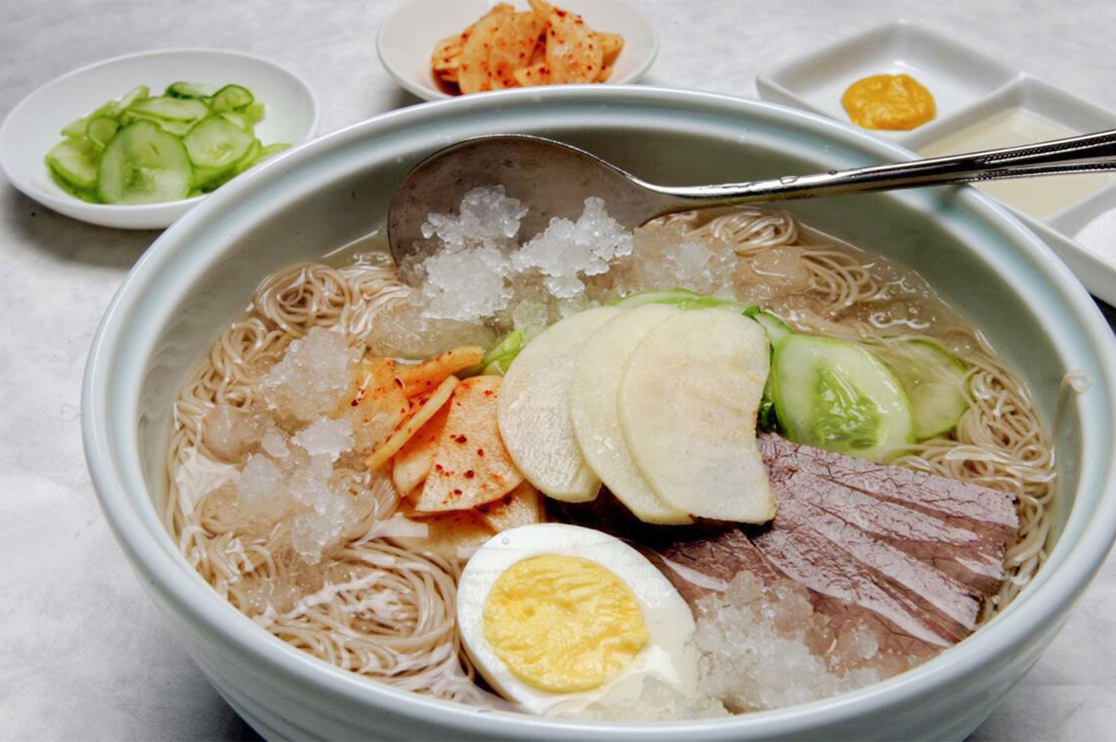 Bowl of Mul Naengmyeon, Korean cold buckwheat noodles served in an icy broth topped with sliced Asian pear, cucumber, pickled radish, beef brisket, and a boiled egg.