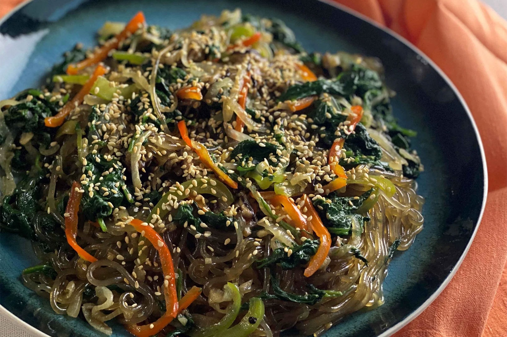 Plate of Japchae, savory Korean sweet potato starch glass noodles stir-fried with spinach, carrots, bell peppers, onions, and topped with sesame seeds.