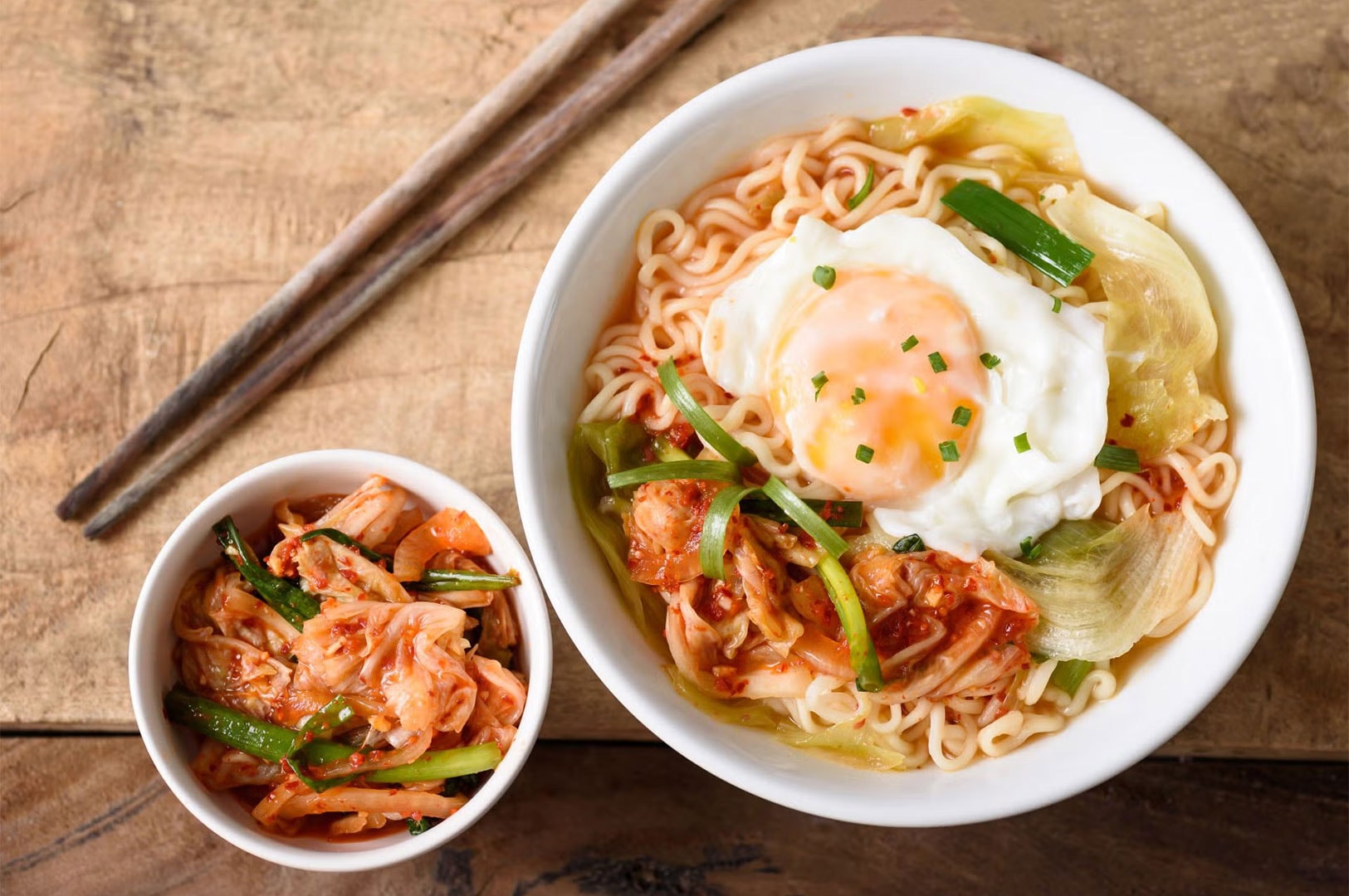 Bowl of spicy Korean Ramyeon noodles topped with a fried egg and scallions, served alongside a small bowl of spicy cabbage Kimchi.