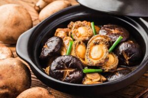 Close-up of braised whole abalone and shiitake mushrooms garnished with green onions served in a traditional black clay pot.