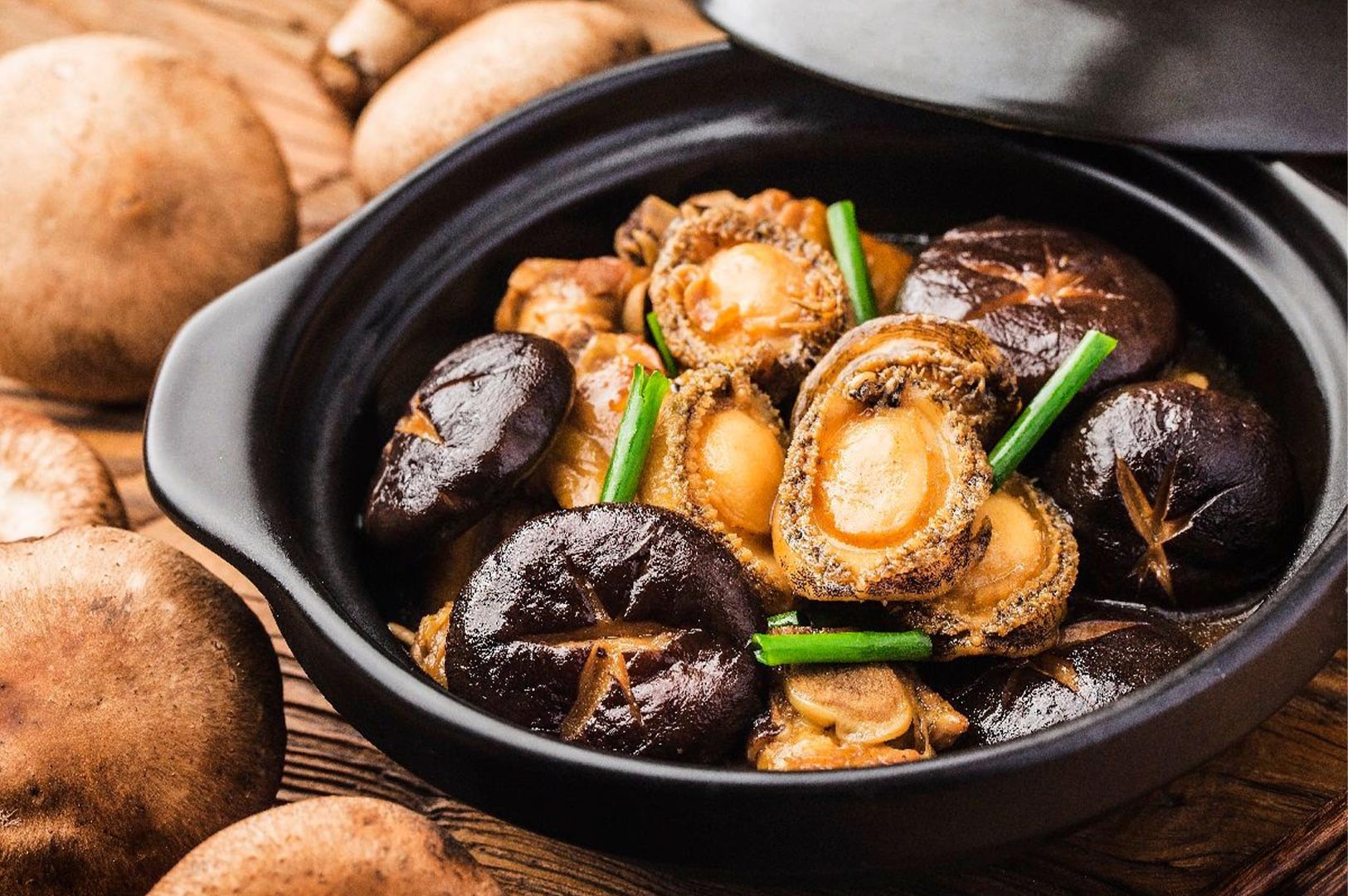 Close-up of braised whole abalone and shiitake mushrooms garnished with green onions served in a traditional black clay pot.