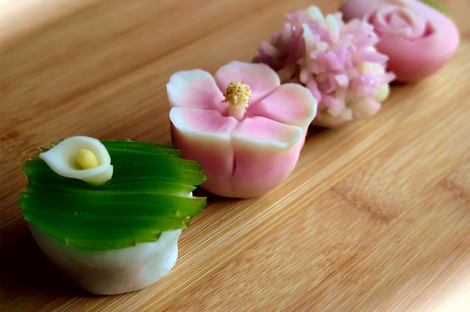 A row of four intricate, floral-shaped Wagashi (traditional Japanese sweets) in pink and green colors displayed on a bamboo serving tray.