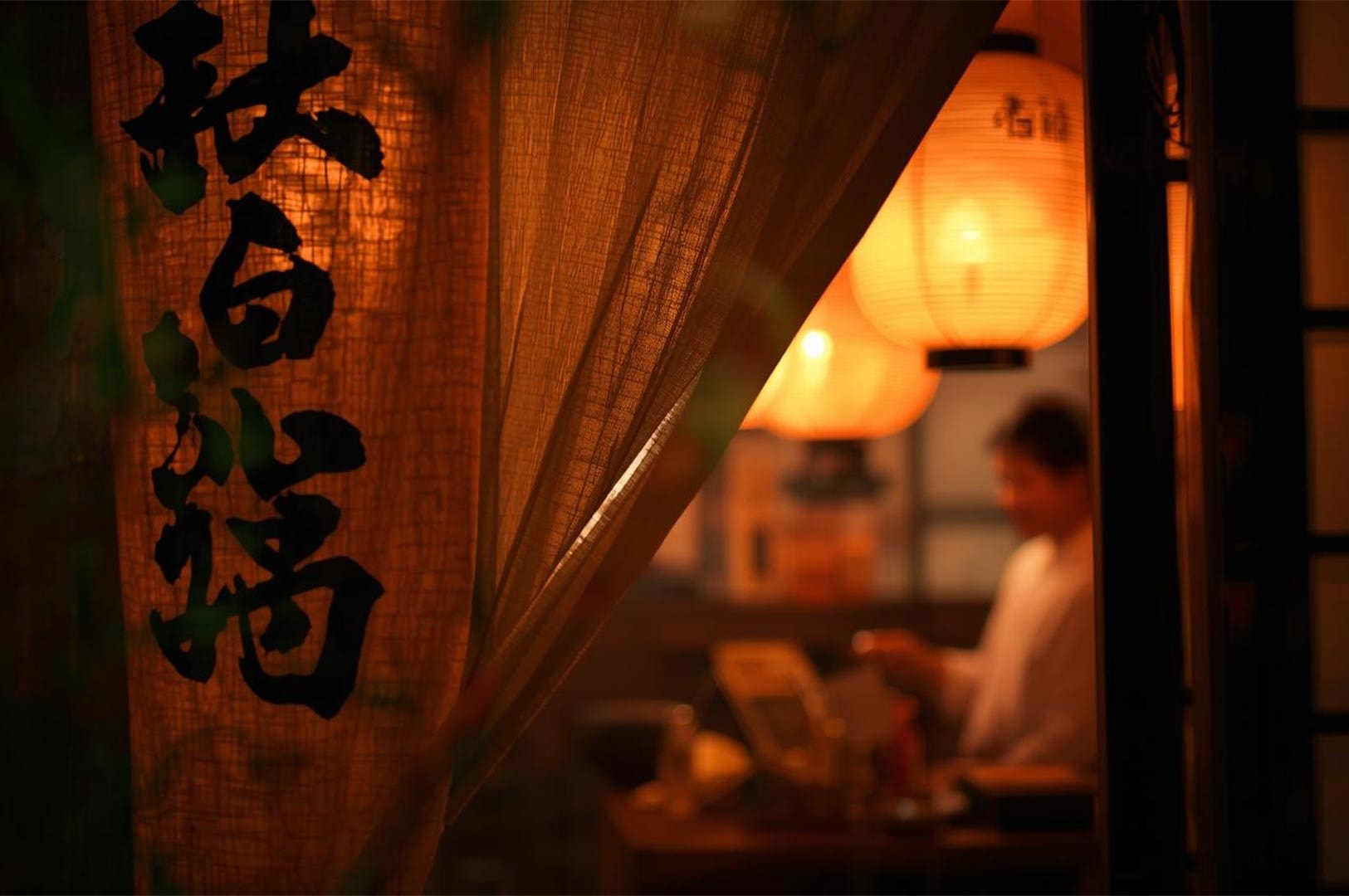 A warm, inviting view of a Japanese Izakaya entrance featuring glowing red paper lanterns and a traditional noren curtain with calligraphy.