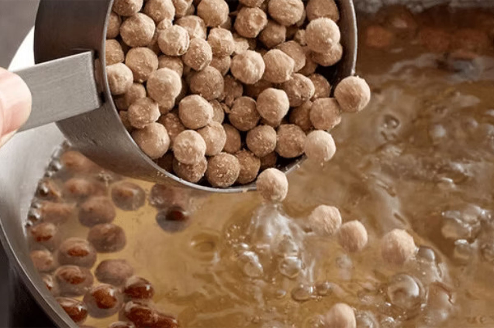 Dry, uncooked brown tapioca pearls being poured from a metal measuring cup into a pot of boiling water to make boba.