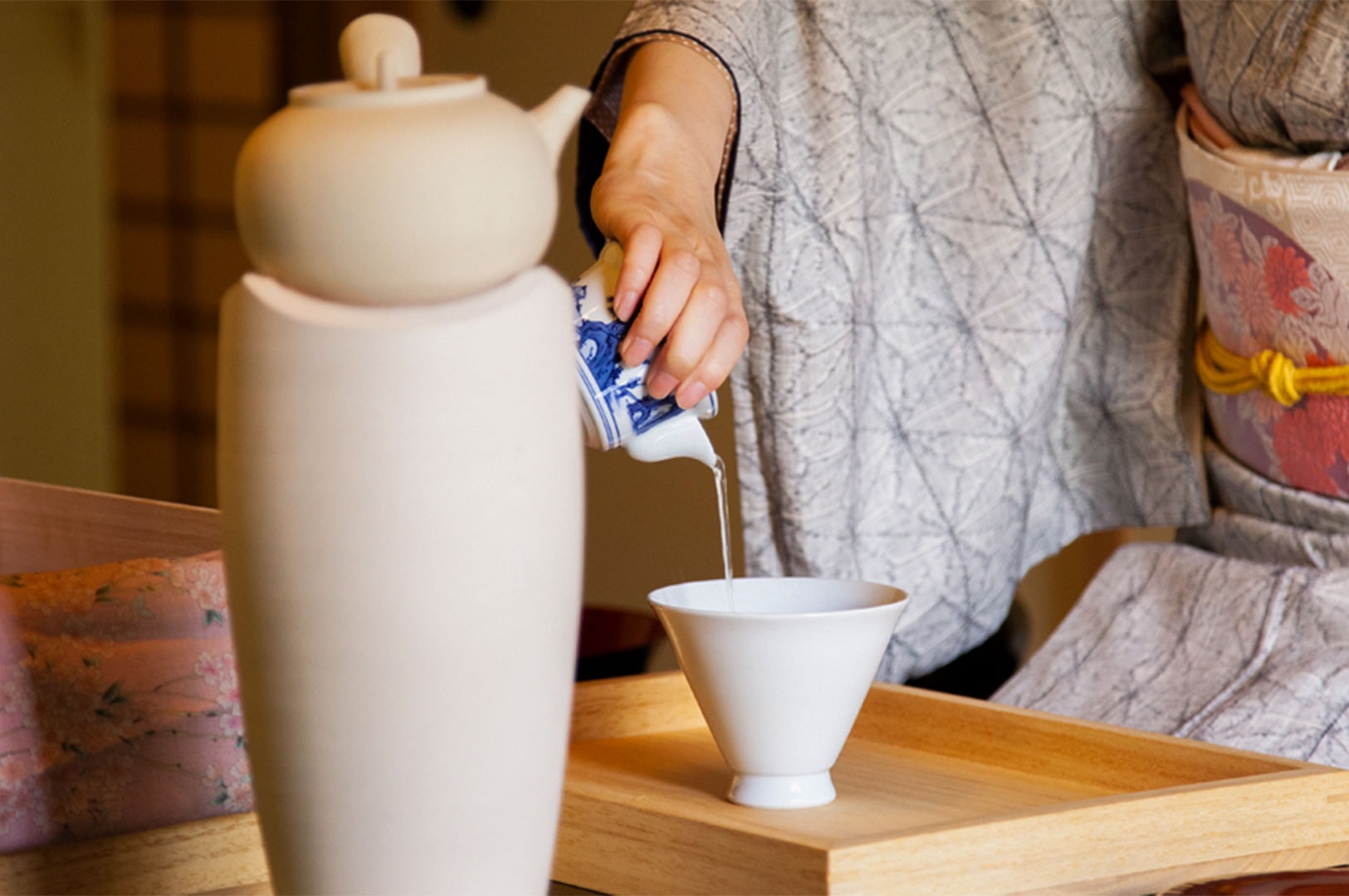 A person wearing a traditional patterned kimono pouring hot water from a pitcher into a white bowl during a formal Japanese tea ceremony.