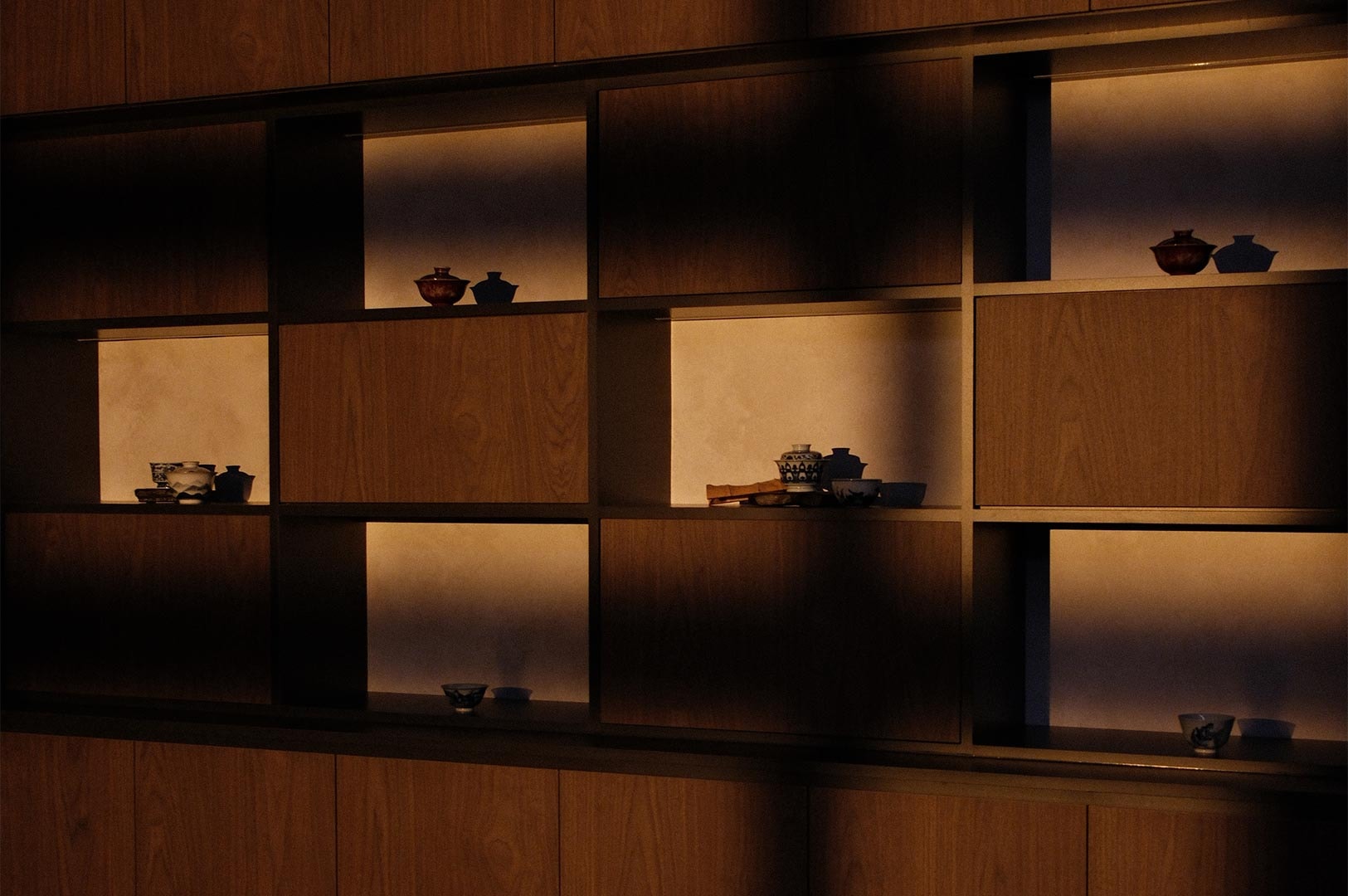 A wide shot of a backlit wooden shelving unit displaying a curated collection of antique and contemporary ceramic teaware.