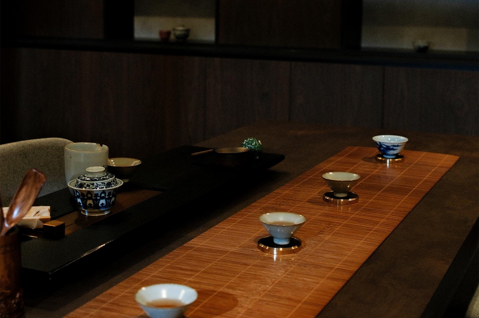 A long wooden tea table set for a ceremony with a bamboo runner, multiple tasting cups, and traditional brewing vessels in a Zen-style room.
