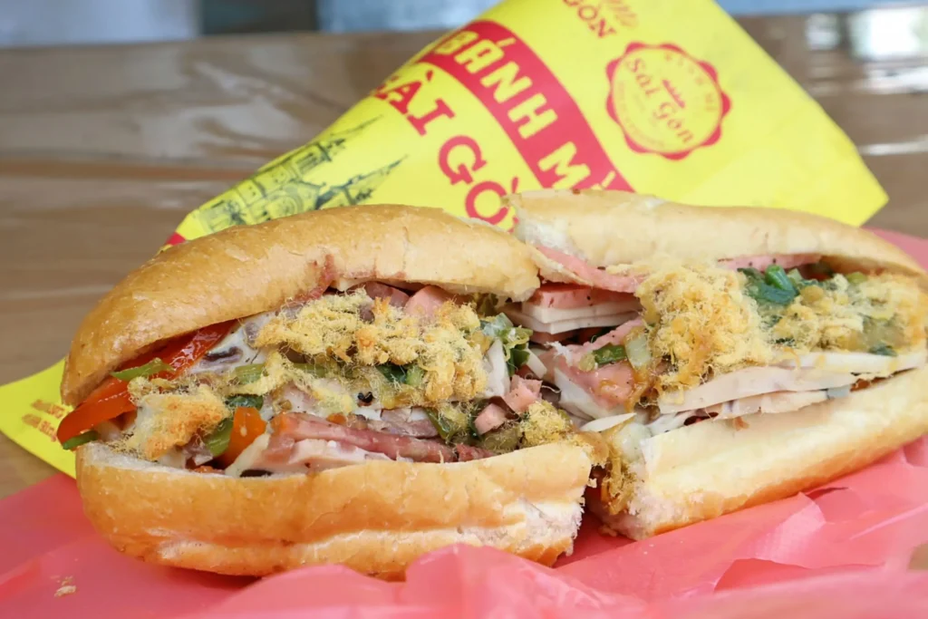 The image features a crusty baguette sandwich filled with various meats, herbs, and savory pork floss. It is presented on a pink plastic wrapper with a yellow branded paper sleeve visible in the background.