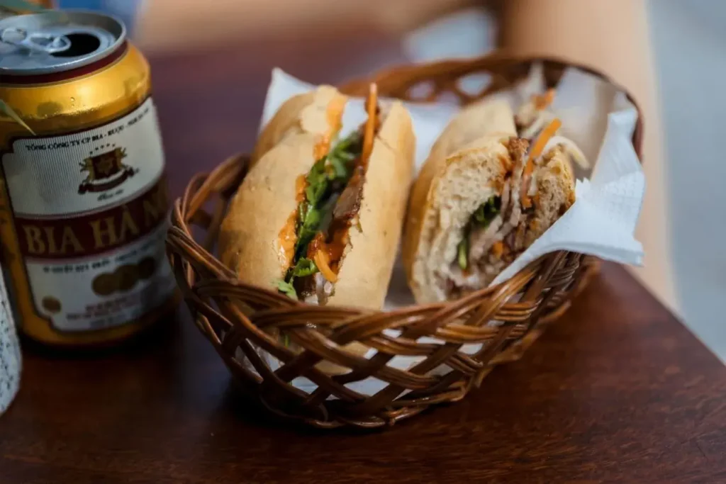 The image features a crusty banh mi sandwich filled with meat and fresh herbs served in a small woven basket. Beside the basket sits a gold-labeled can of Bia Hanoi beer on a dark wooden tabletop.