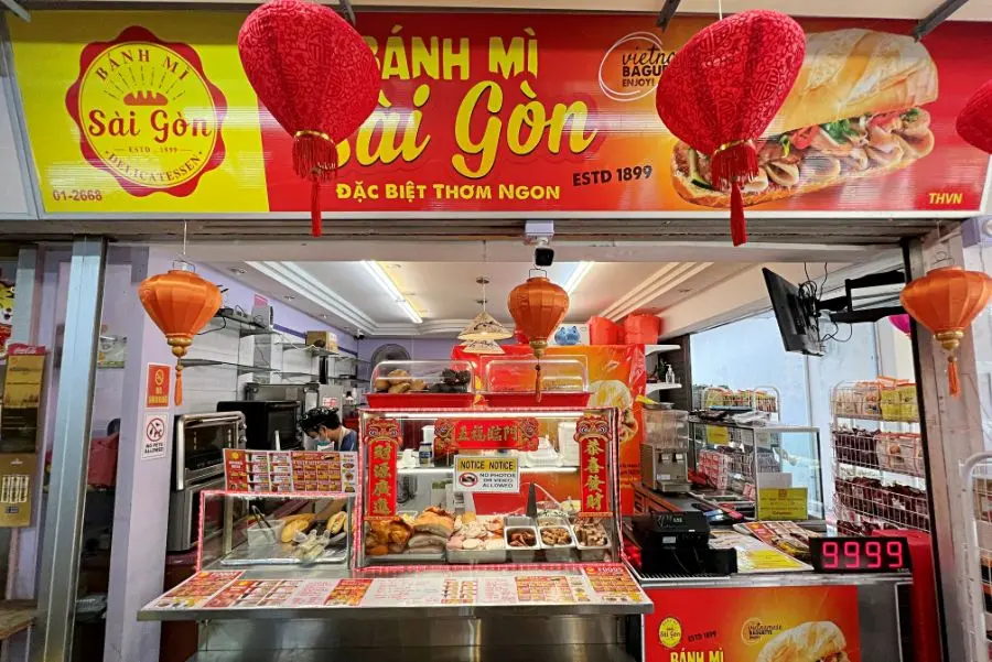 The image shows the vibrant storefront of a Vietnamese Bánh Mì shop decorated with traditional red and orange lanterns. A glass display case at the counter showcases various fillings and ingredients used to prepare the sandwiches.