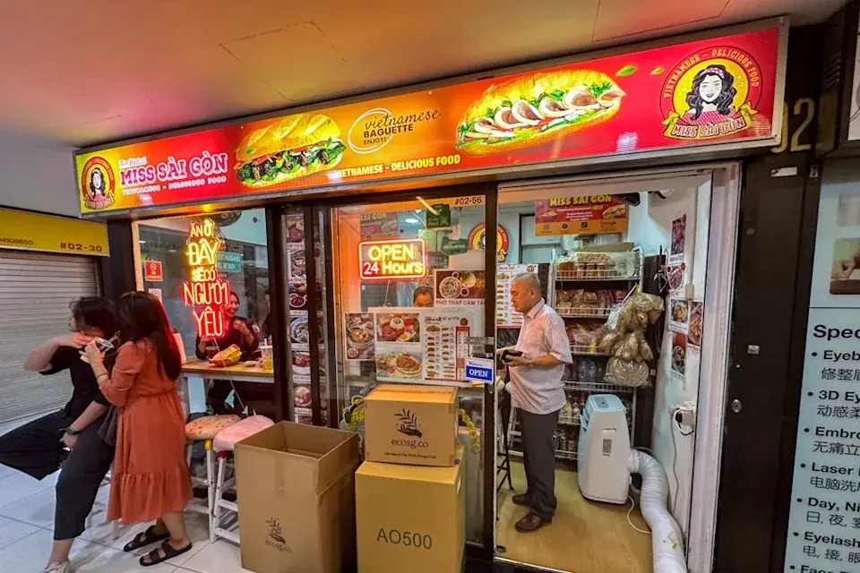 The image shows the storefront of a small Vietnamese restaurant called "Miss Sài Gòn" that is open 24 hours. A man stands near the entrance next to cardboard boxes, while a group of people sits at a high table visible through the window.