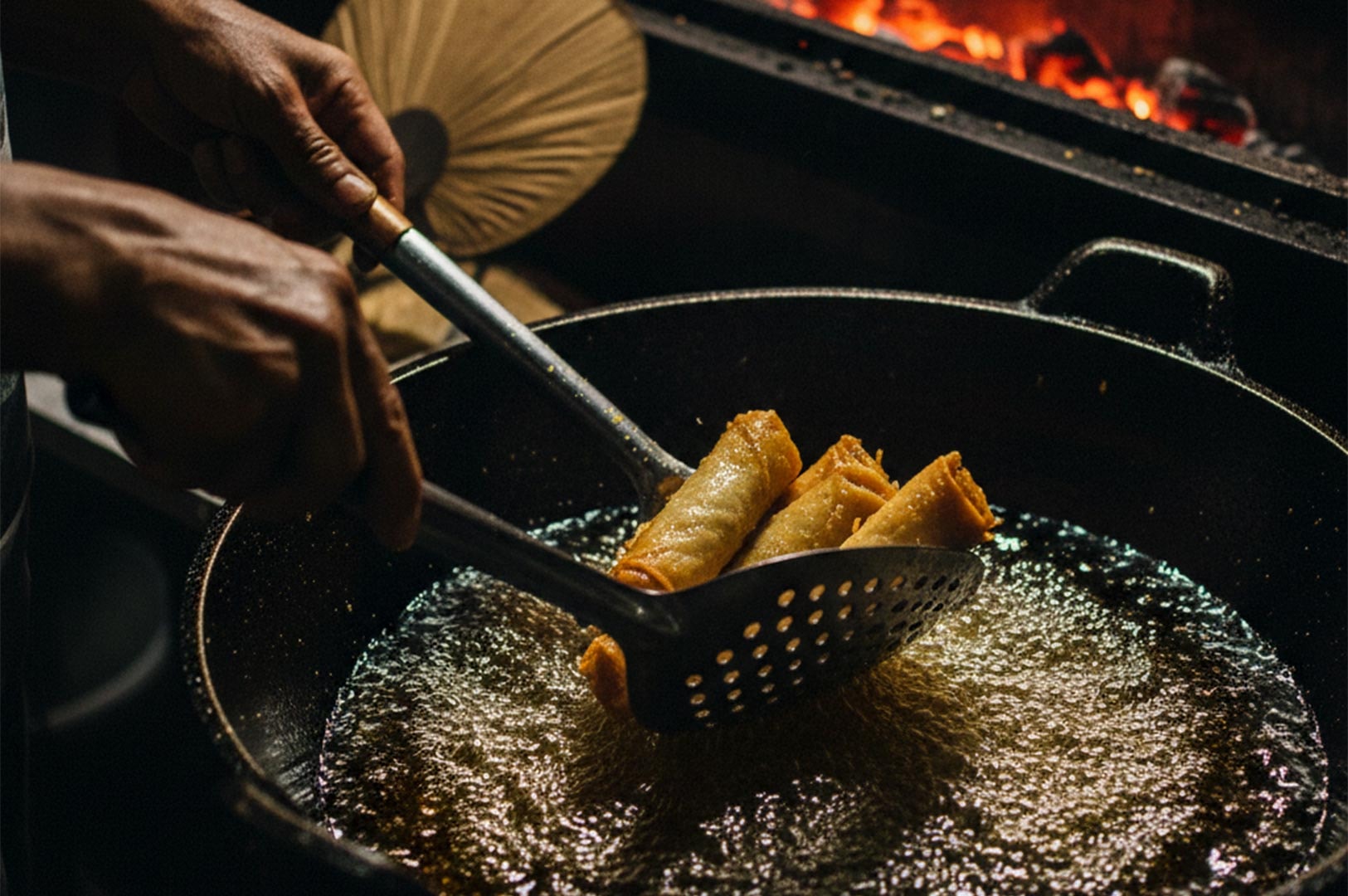 A close-up shot of a street food vendor using a slotted metal spatula to scoop crispy fried lumpia out of a bubbling wok of hot oil.