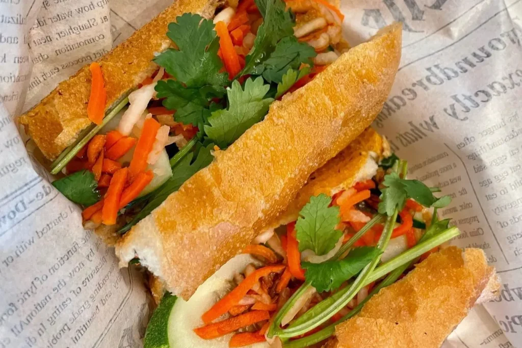 The file shows a Vietnamese banh mi sandwich served in two halves on newsprint-patterned paper. Each section of the crusty baguette is generously filled with fresh cilantro, pickled carrots, and cucumber slices.