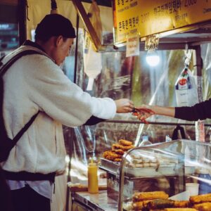 A customer in a white fleece jacket pays a street food vendor at a small night market stall displaying fried snacks under warm lighting.