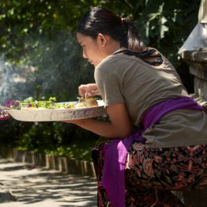 A woman in traditional attire kneels outdoors beside a stone shrine, holding a tray with flowers and incense as smoke rises in a quiet, leafy setting.