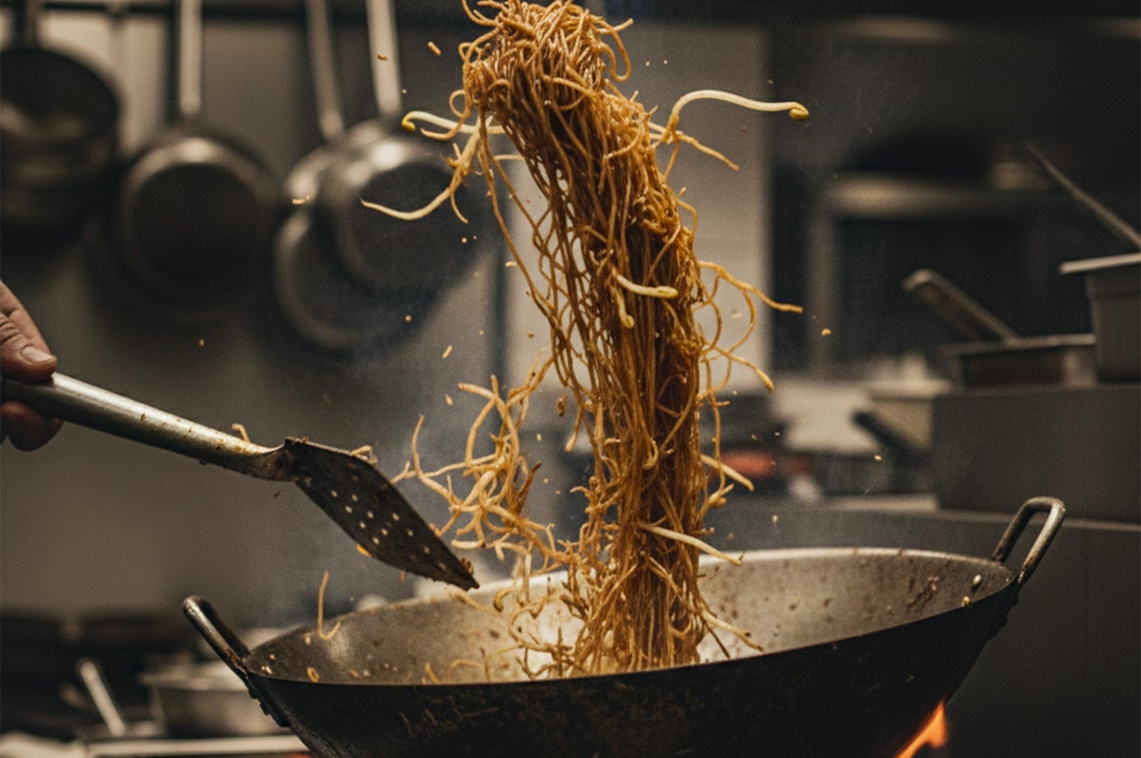 Action shot of a chef tossing long noodles and bean sprouts in a large metal wok over a high flame in a professional kitchen.