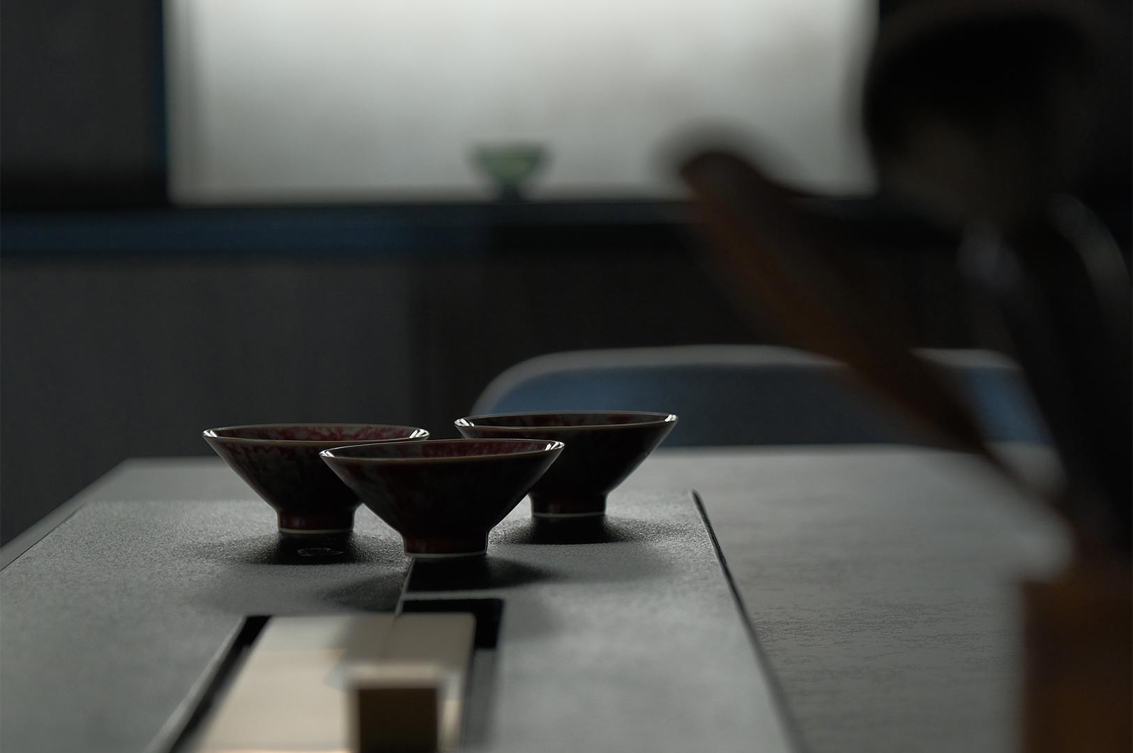 Three small dark red porcelain tea cups resting on a black textured tea tray in a softly lit, moody setting.