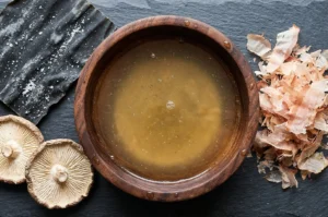 Top-down view of a wooden bowl of clear dashi stock surrounded by dried kombu, shiitake mushrooms, and bonito flakes.