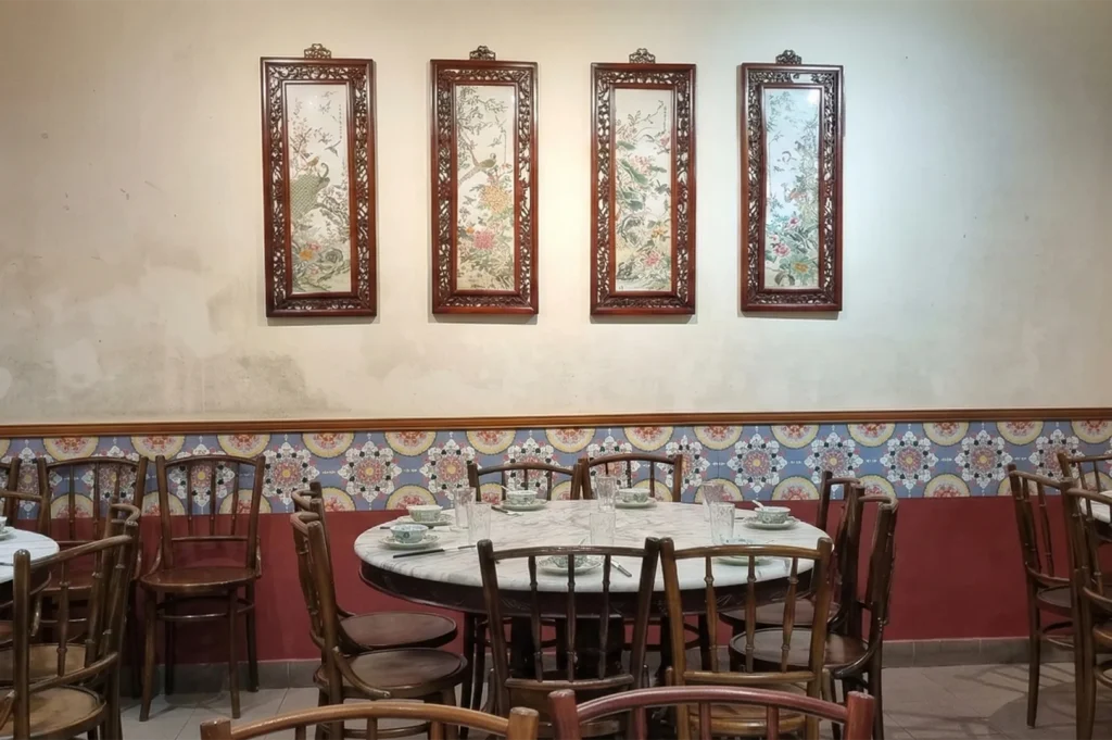 Interior of a traditional Chinese restaurant featuring marble-top round tables, wooden chairs, and four large framed silk paintings on a cream wall above decorative Peranakan tiles.