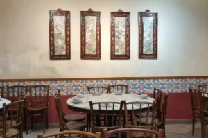 Interior of a traditional Chinese restaurant featuring marble-top round tables, wooden chairs, and four large framed silk paintings on a cream wall above decorative Peranakan tiles.