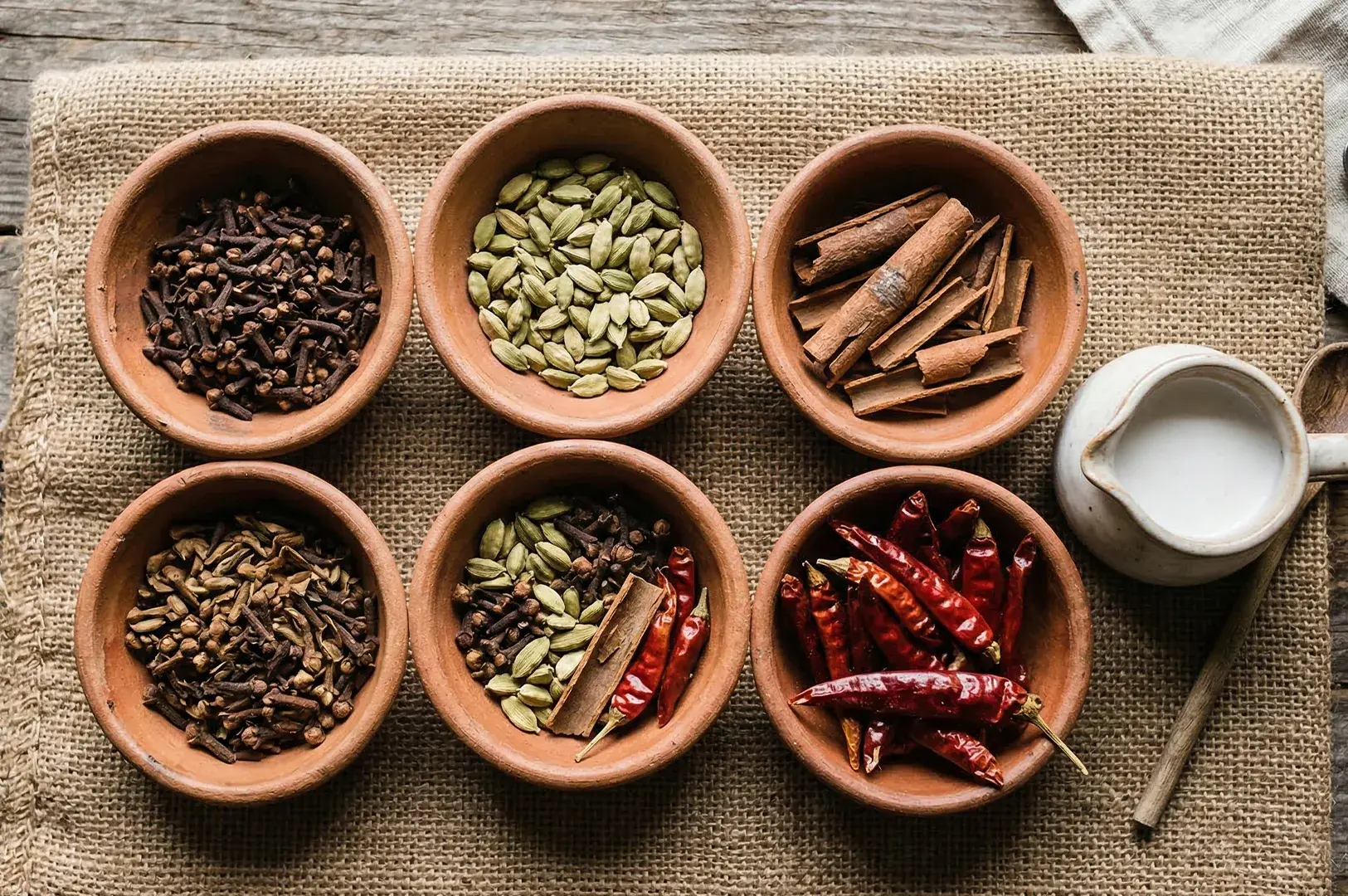 A top-down view of six clay bowls containing various whole spices including cinnamon sticks, green cardamom, dried chilies, and cloves.