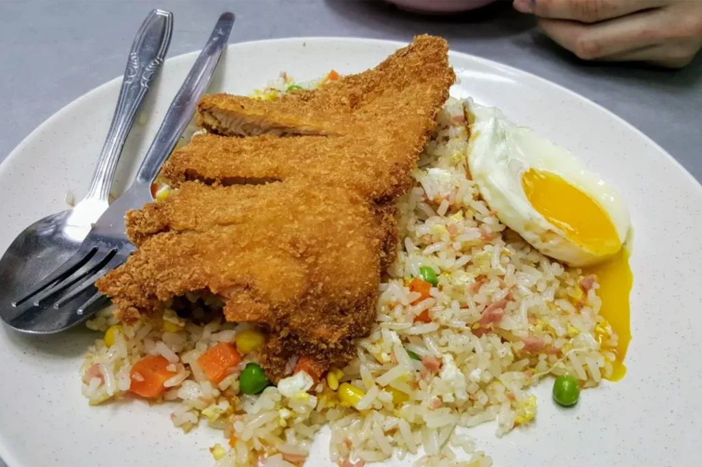 Crispy golden-brown chicken cutlet sliced over a bed of egg fried rice with peas, corn, and carrots. A runny sunny-side-up egg sits on the side, served on a classic speckled hawker plate with silver cutlery.