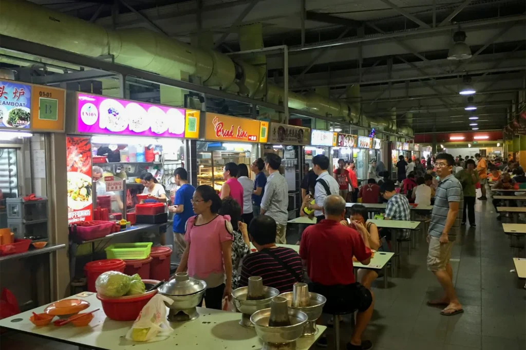 A busy indoor Singaporean hawker centre with various food stalls like Bread Line and Steam Boat, featuring locals dining at communal tables.