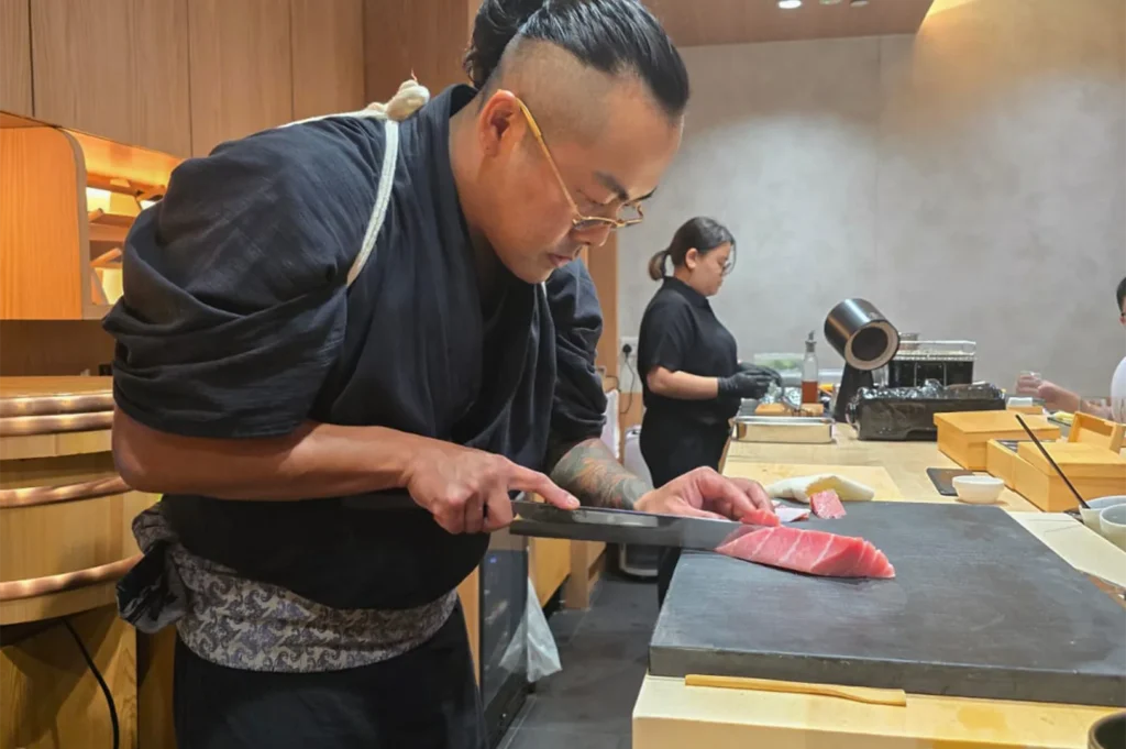 Expert sushi chef in a black kimono slicing a large fillet of fresh bluefin tuna on a professional cutting board.