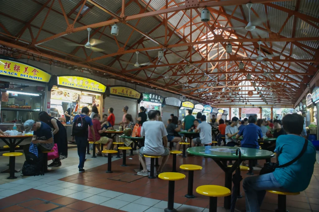 A wide shot of a bustling Singaporean hawker center with people sitting at round tables and various food stalls like "Rojak" and "Peanut Soup" under an orange industrial roof.