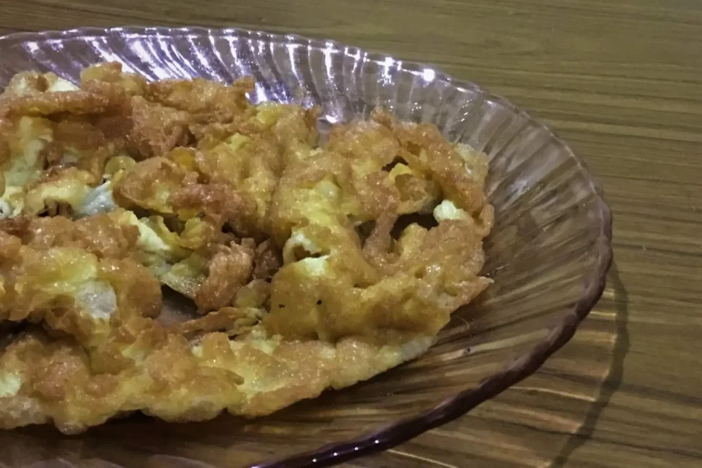A golden-brown Indonesian fried omelet, known as Telur Dadar, rests on a pink glass plate. The eggs have a distinctively crisp, lacy texture from being deep-fried in hot oil.