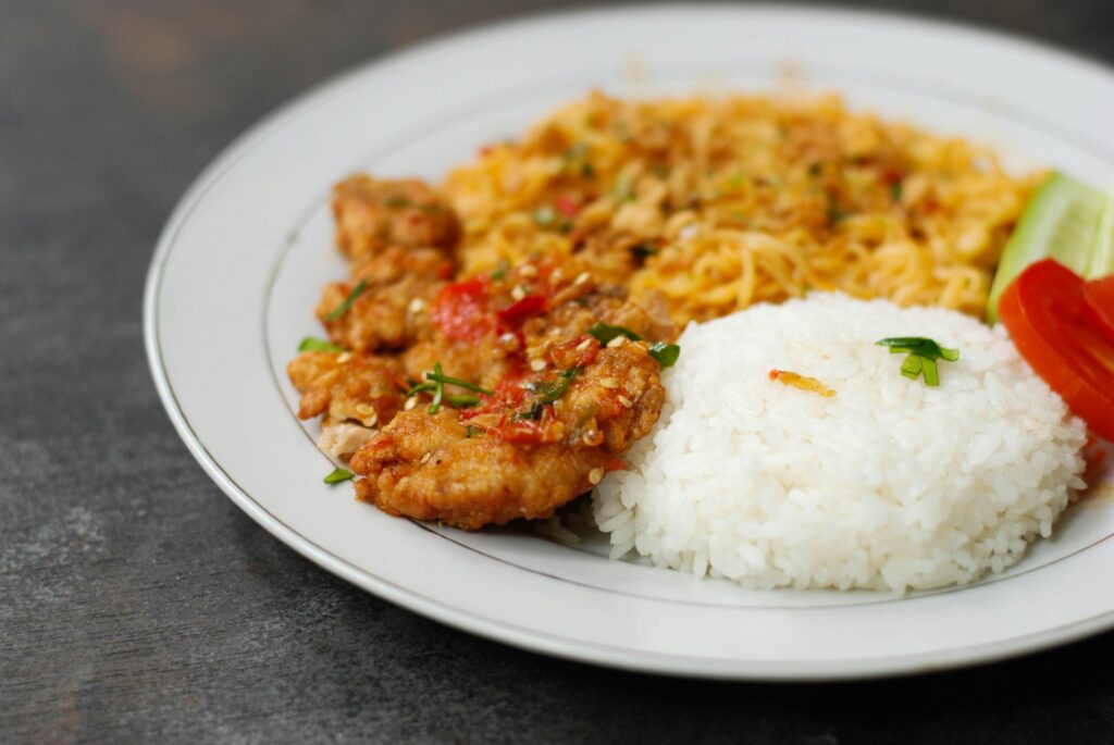 This image features a white plate topped with a mound of steamed white rice, crispy fried chicken with chili sambal, and a side of stir-fried noodles. Slices of fresh cucumber and tomato garnish the edge, providing a bright contrast to the savory elements on the dark textured surface.