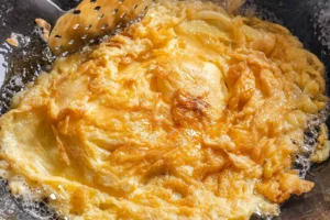 A golden-brown Thai-style omelet sizzles in a wok filled with hot, bubbling oil. A metal slotted turner rests near the edge, ready to flip the crispy, aerated eggs.