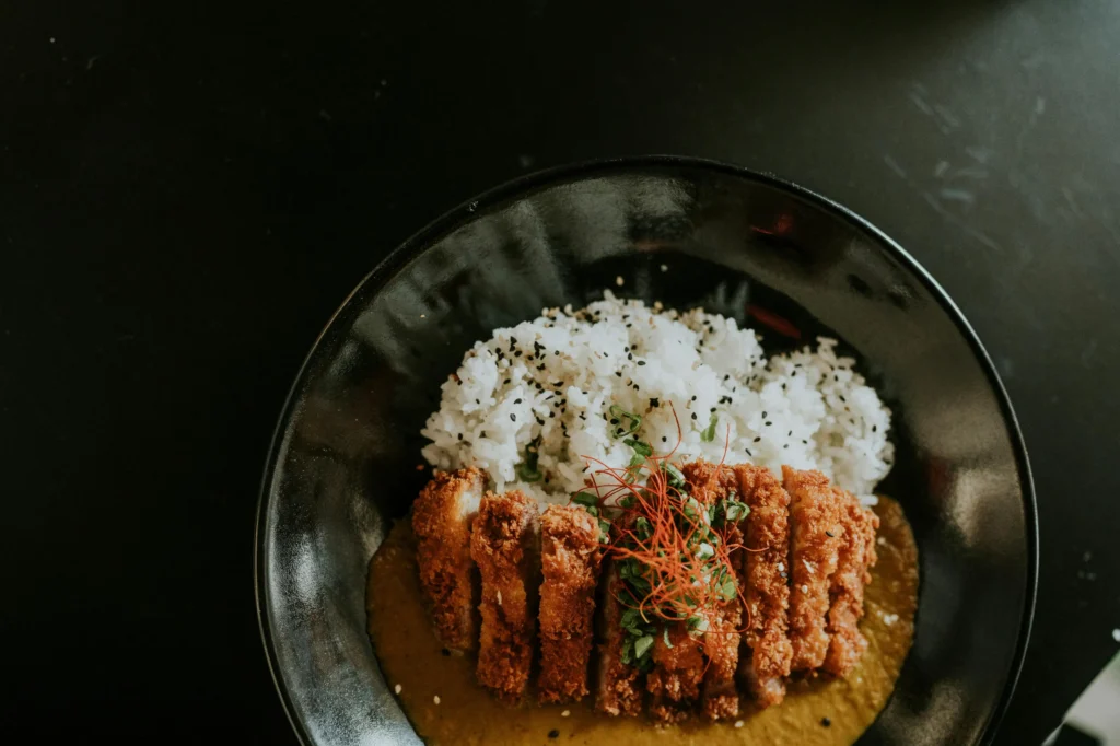 This high-angle shot captures a bowl of Japanese chicken katsu curry served on a dark, minimalist table. A golden-brown breaded cutlet is sliced and plated over a rich curry sauce, accompanied by a bed of white rice garnished with black sesame seeds and vibrant chili threads.