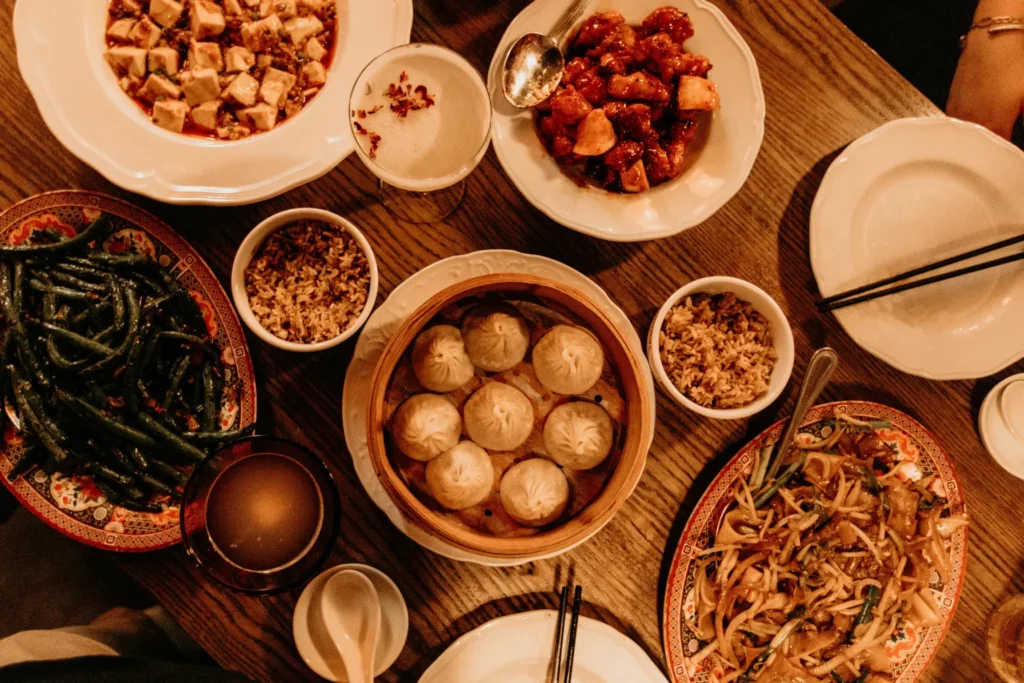 A wooden table is crowded with an array of Chinese dishes, including a steamer basket of soup dumplings, mapo tofu, stir-fried green beans, and noodles. The scene is captured in warm, dim lighting, featuring several plates of food, bowls of rice, and a floral-garnished cocktail.