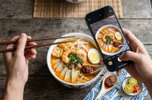 A top-down view of a hand holding a smartphone taking a photo of a steaming bowl of Laksa with prawns, egg, and fish cakes on a rustic wooden table.